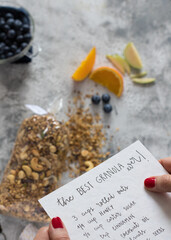 Closeup view of a granola recipe held by both woman hands written with lettering over some granola...