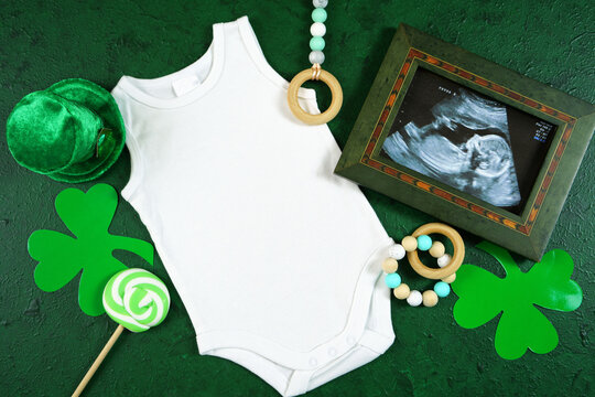Happy St Patrick's Day baby wear sleeveless onesie bodysuit and sonogram, styled with leprechaun hat, shamrocks, on a textured green background. Mockup. Top view flat lay. Copy space.