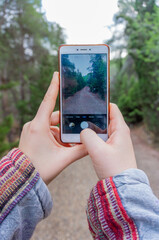 Vertical shot of a female hands holding a mobile phone while take a photo of a path on the forest to share it in social media