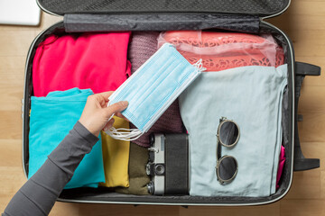 A woman's hand puts a mask into an open suitcase packed with travel stuff.