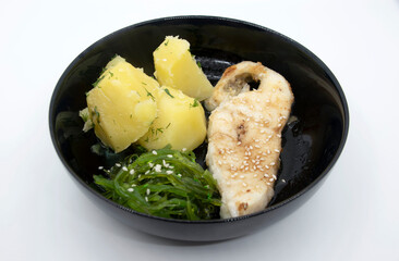 Fried sea fish steak boiled potatoes Chuka seaweed salad on a black plate