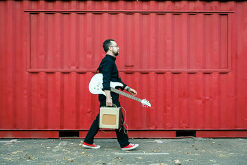 Man in black with a beard with electric guitar walking by the red container