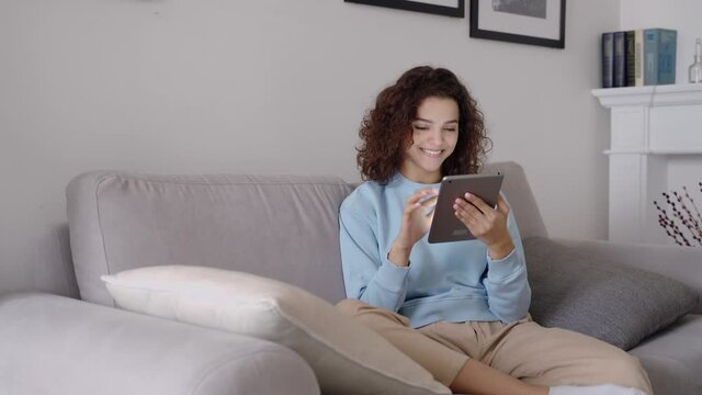 Happy hispanic teen girl holding pad computer gadget using digital tablet technology sitting on couch at home. Smiling young woman using apps, shopping online, reading news, browsing internet on sofa.