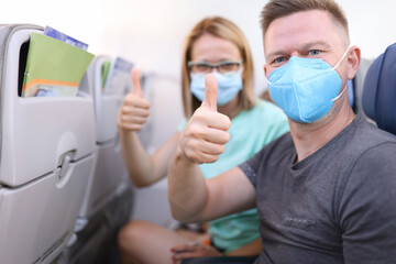 Fototapeta na wymiar Man and woman in medical protective masks in aircraft cabin hold their thumbs up. Safe flights to coronavirus pandemic concept