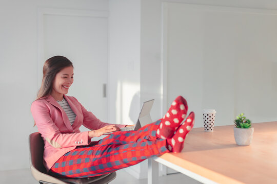 Work online remote from home funny concept. Asian woman relaxing in pajama pants and cozy socks while wearing professional top and suit for videocall meeting.