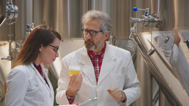 Food scientists in white coats discussing a beer sample and taking notes. Two food technologists with glasses talking about a beer sample. Science, brewery concept.