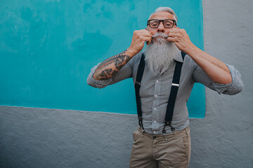 an older man in hipster clothes and glasses and a long white beard poses on a blue wall