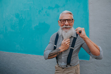an older man in hipster clothes and glasses and a long white beard poses on a blue wall