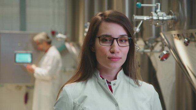 Portrait of a female scientist smiling and looking at camera. Female scientist in a white lab coat wearing glasses. Male scientist working in the background. Science concept.
