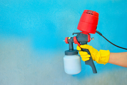 A worker's hand in a yellow glove holds a hand-held spray gun with an overhead compressor. Painting the wall of the house in blue. Household renovation. Copy space. Selective focus