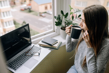 Young woman drinking coffee relaxing from work, talking on the phone. Workplace on the windowsill at home.