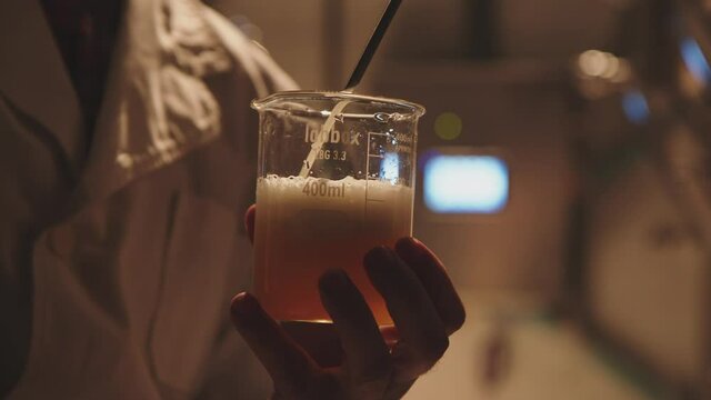 Close-up of a hand of a scientist holding beaker and pouring beer into it for testing. A worker in a white lab coat. Beer foam. Science, brewery concept.