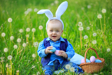 Adorable toddler boy wearing bunny ears playing with Easter eggs in basket sitting in sunny garden with white spring flowers. Smiling baby having fun outdoors in park. Copy space. Happy Easter holiday