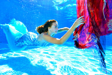 A young girl in a shiny blue dress swims underwater in a pool and catches a red cloth that sinks in the water. Portrait. Side view. Underwater photography. Horizontal orientation.