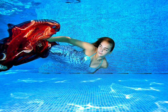 A young girl swims underwater in an outdoor pool in a shiny blue dress and with a red cloth in her hands on a bright sunny day. Portrait. Underwater photography. Side view. Horizontal orientation.