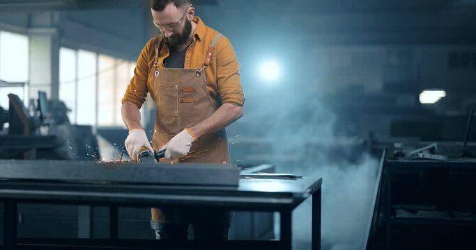 Metal industry worker dressed in apron doing some metalwork, grinding steel product at the factory. Handsome artisan at the manufacturing of metal products. Tilt shot