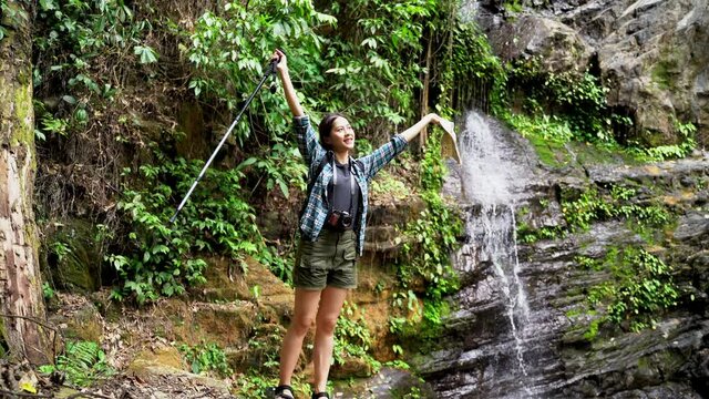 Asian female traveler traveling hiking adventure exploring nature jungle trail into forest waterfall, wearing travel gear coat, bag, and walking stick