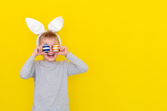 Happy Easter kids. Boy in rabbit bunny ears on head with colored eggs on yellow studio background Cheerful crazy smiling child copyspace