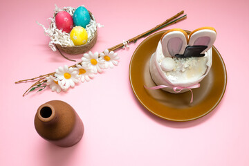 White cake with bunny ears and black hat in brown plate, three colorful eggs in nest, brown bottle on the white and pink background. Composition decorated with flowers