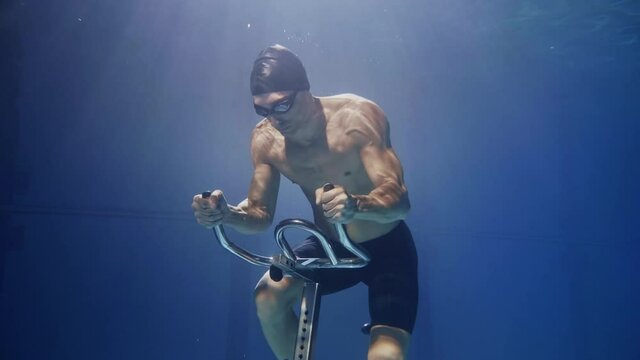 Cinematic shot of young male athlete with muscular body is exercising with effort and determination on stationary cycling machine underwater. Concept of sport, fitness, healthy lifestyle, recreation.