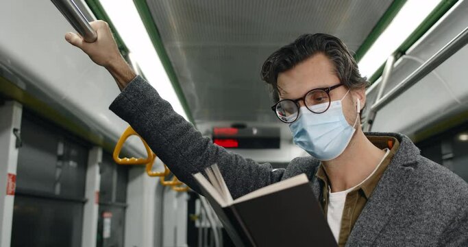Crop view of man in medical mask holding book in tram. Male person in glasses and wireless earbuds reading story and holding handrail in public transport. Concept of commuting.