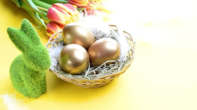 Golden eggs in basket with spring tulips, white feathers on pastel yellow background in Happy Easter decoration. Foil minimalist egg design, modern design
