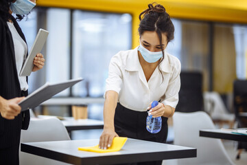 Young businesswomen clean the work place, wipe the desk with yellow rag. Colleagues disinfect the working surface with a sanitizer spray to stop covid-19 spread. New normal, office hygiene concept.