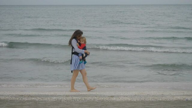 Woman hold child in baby wearing walk on beach coast, peaceful holiday, sea wave