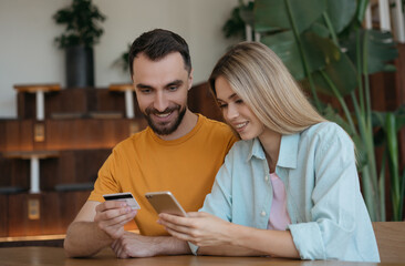 Smiling couple ordering food sitting at home, delivery concept. Happy friends holding credit card, using mobile phone, shopping online