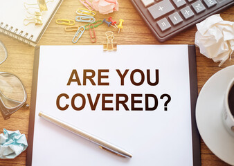 On a wooden table there is an office sheet of paper with the text ARE YOU COVERED. Business workspace with calculator, glasses, pen, crumpled paper and cup of coffee.