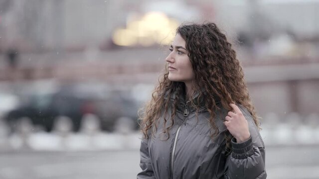 A young woman with curly hair poses on a city street, wearing a jacket, cold season. She walks.