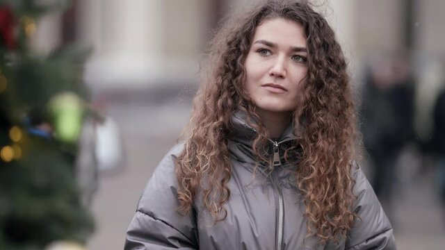 Close-up portrait of a young woman with curly hair posing on a city street, wearing a jacket, cold season. She poses near the Christmas tree.