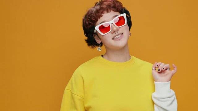 Dancing Girl Teenager Asian Listens to Music in Headphones from Smartphone on Yellow Background with Piercings in Glasses. Smiling with Bright Makeup moving Up Hands Slow Motion. Positive emotions