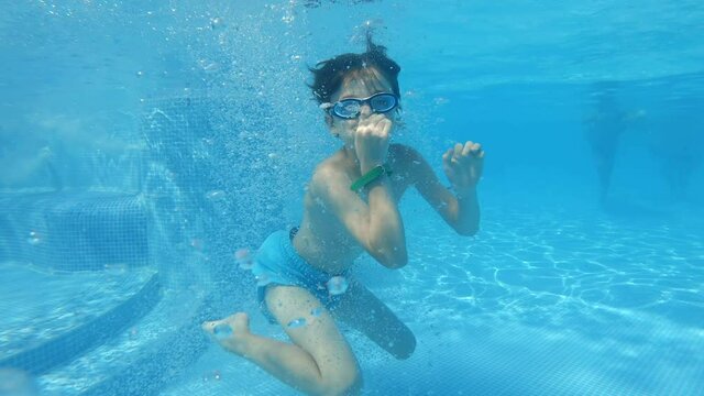 Happy boy jumping into a pool splashing in all directions and swimming under blue water smiling slow motion in a summer water park on a sunny day on vacation with his family. Travel. Tourism