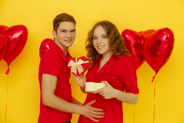 Birthday holiday party concept. Beautiful girl dressed in red dress opens gift box. Young loving couple in red clothes celebrating Valentines Day isolated on yellow background with red balloons.