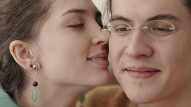 Close up of lovely young Caucasian woman wearing beautiful earrings kissing her beloved boyfriend wearing glasses on cheek