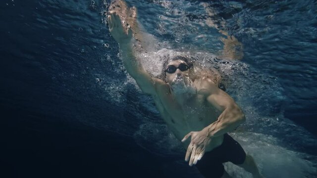 Cinematic shot of young professional male swimmer with cap and goggles trains with effort and dedication to win the race by practicing in swimming pool. Concept of sport, competition, victory, fitness