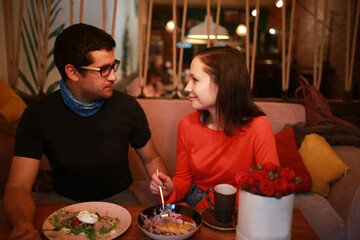 The young couple have breakfast in a cafe, they celebrate their anniversary and Valentine's Day, they have a date. A man with glasses gave his girlfriend red roses in a box. They drink coffee or tea. 