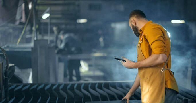 Factory worker supervising production of metal goods with a digital tablet at the factory. Supervision of heavy industry