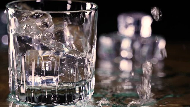 Close up of ice cubes dropping into glass of water splashing onto wood counter in slow motion