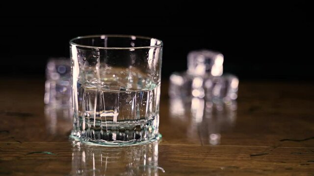 Ice cubes spin and ricohete violently as they are dropped in a glass of water on wood counter. In slow motion