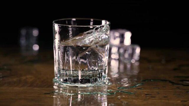 Slow zoom in of ice cubes dropping into glass of water splashing onto wood counter in slow motion