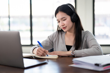 Young asian girl student wears wireless headphones write on the notebook to study language online watch and listen to the lecturer, webinar via video call e-learning at home, distance education