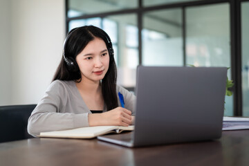 Young asian girl student wears wireless headphones write on the notebook to study language online watch and listen to the lecturer, webinar via video call e-learning at home, distance education