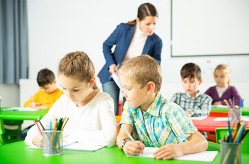 Fototapeta premium Assiduous school kids with pens and notebooks studying in classroom with female teacher