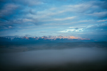 Scenic mountain landscape with great snowy mountain range lit by orange dawn sun above dense fog among low clouds at twilight. Awesome morning scenery with high mountain ridge under blue cloudy sky.