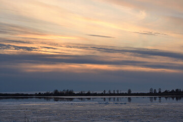 A landscape view of the frozen sea during ice drift in spring with dramatic sky and with reflections in melt water in the evening at sunset. In the distance, the black strip of the island.