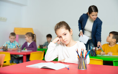 Upset small girl sitting at table in schoolroom on background with pupils studying with teacher