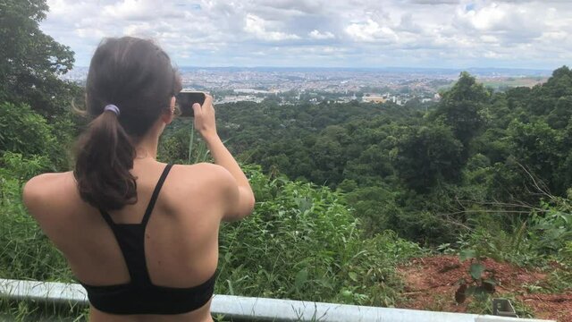 Rear View Of Female Tourist-Hiker Taking A Panoramic Picture Of The Cityscape Using Her Smartphone On Mountain Forest On A Sunny Day In Praia Porto, Brazil - Medium Closeup Shot