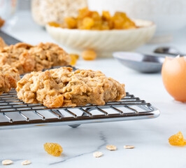 Close up of oatmeal raisin cookies cooling on a metal cooling rack with ingredients in soft focus in behind.